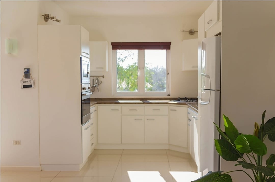 A modern kitchen is featured with white cabinetry and a variety of appliances, including an oven and refrigerator. A window above the sink allows natural light, and a green plant adds a touch of color. Clean lines and a spacious layout enhance the cooking area.