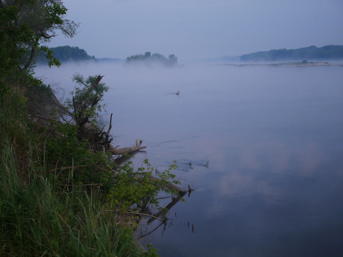 A serene river scene is captured at dawn, shrouded in a light mist. The still water reflects the soft hues of the morning sky, while lush greenery lines the riverbank. Gentle contours of the landscape emerge in the distance, creating a tranquil ambiance.