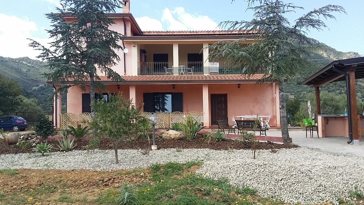 A spacious, two-story house features a light-colored facade with a covered porch and balcony. The surrounding landscape includes landscaped garden areas with gravel, shrubs, and trees. An outdoor dining area is visible, along with a barbecue space nearby, providing a welcoming atmosphere.
