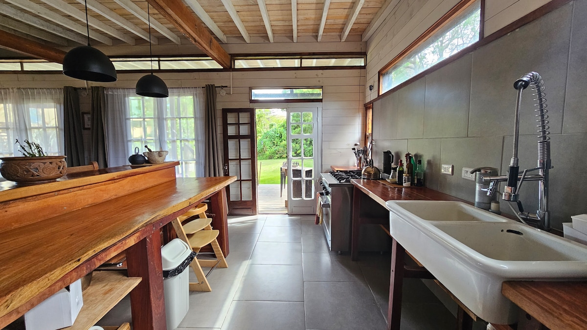 A well-equipped kitchen features wooden countertops and open shelving. Natural light filters through large windows, illuminating the space. A dining area with simple chairs is visible, alongside modern appliances and a stylish sink, enhancing the functional layout.