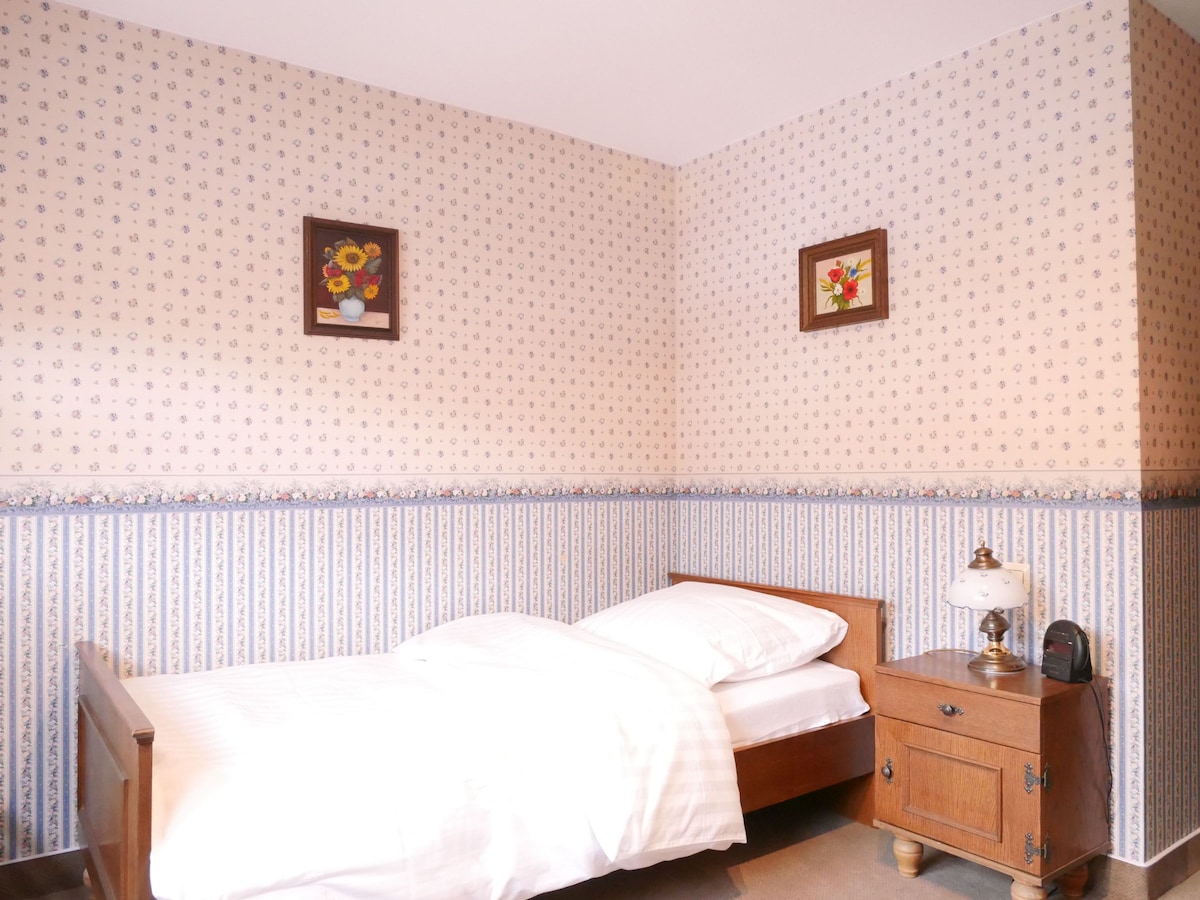 A cozy bedroom features a single bed with crisp white linens. Floral-patterned wallpaper adorns the walls, complemented by two framed floral artworks. A nightstand holds a vintage lamp and a small clock, creating a simple yet charming atmosphere.