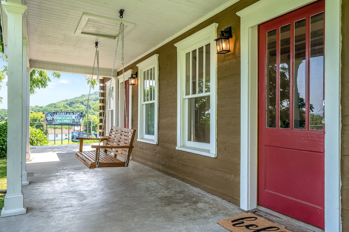 A welcoming front porch features a wooden swing and a bright red door. Large windows offer views of the surrounding greenery, creating a peaceful outdoor space. The floor is covered with a light concrete finish, and a welcome mat adds a touch of hospitality.