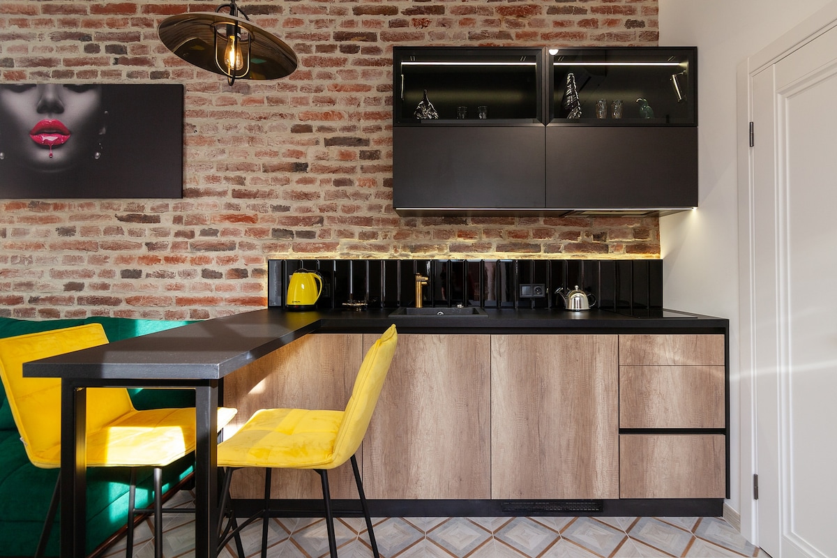 A modern kitchen area features a combination of textured brick walls and sleek cabinetry. A black countertop houses essential appliances, while a small dining table with two bright yellow chairs is positioned nearby, providing a cheerful contrast to the earthy tones.