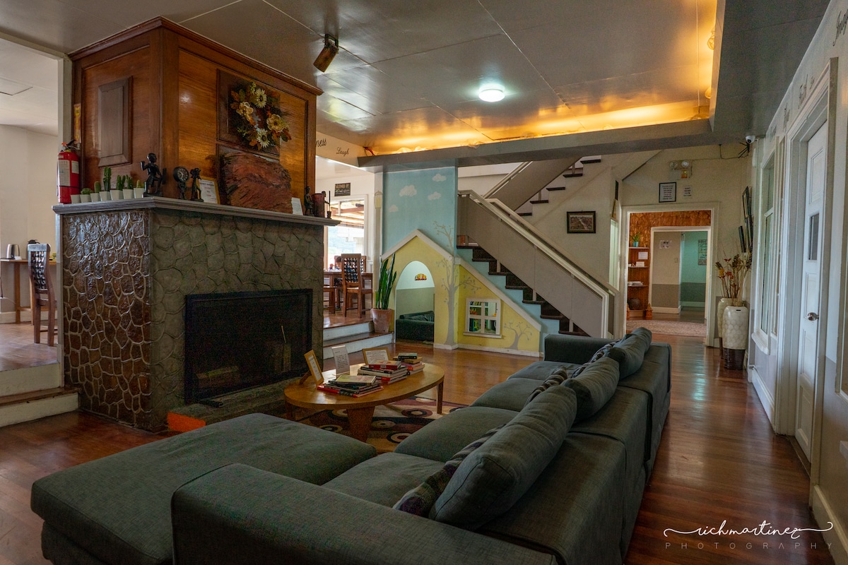 A spacious common area features a stone fireplace and a large, comfortable sofa. The room is bathed in warm light, with a wooden staircase leading to the upper level. Colorful wall art adds character, creating a welcoming and cozy environment.