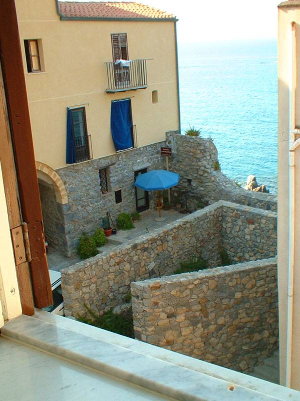 A view from the window showcases a stone courtyard surrounded by weathered walls. A blue umbrella is positioned over a table, providing shade. The serene sea can be seen beyond the courtyard, with gentle waves reflecting the sunlight.