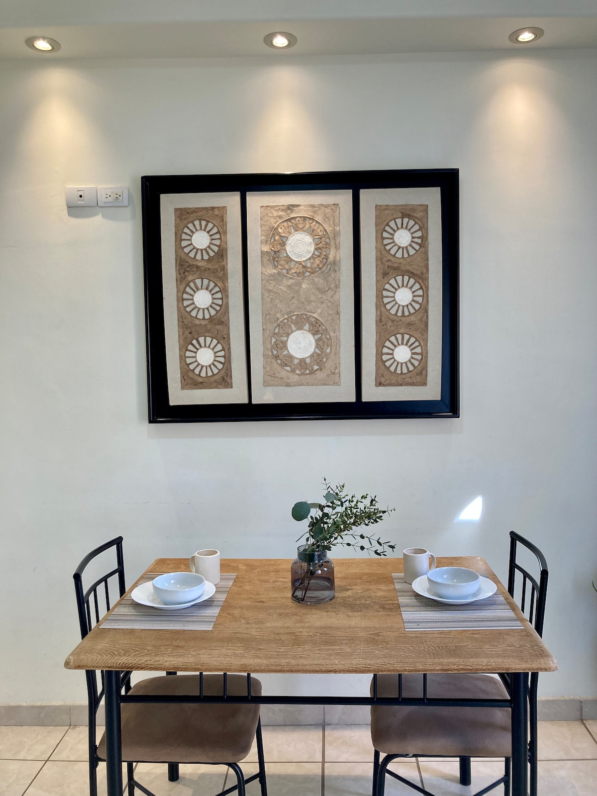 A dining area is presented with a wooden table and four chairs, arranged for two. The table is set with plates and cups, and a small vase containing greenery is centered. A framed artwork is displayed on the wall above, complemented by soft pendant lighting.
