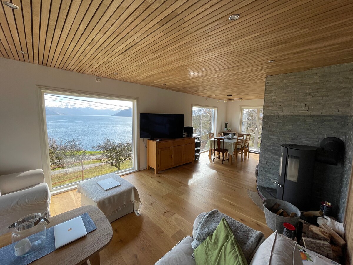 A spacious living area is shown, featuring large windows that frame a view of the fjord and mountains. Natural wood elements are present, with a cozy seating arrangement and a flat-screen TV. A dining table is positioned near the light-filled windows, enhancing the serene atmosphere.