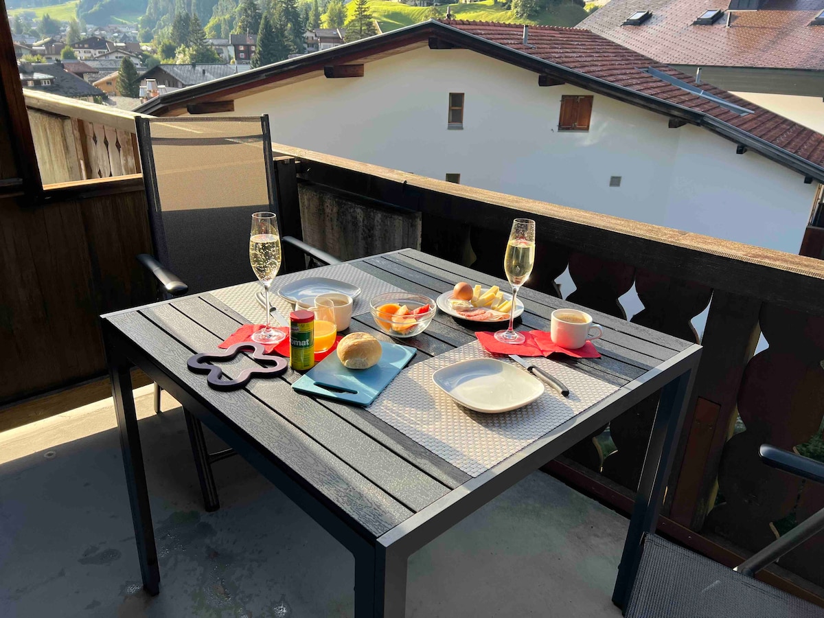 An outdoor balcony table is set for breakfast, featuring two glasses and a variety of dishes, including fruits and pastry. The scenic backdrop of green hills and distant rooftops is visible, providing a pleasant setting for dining.