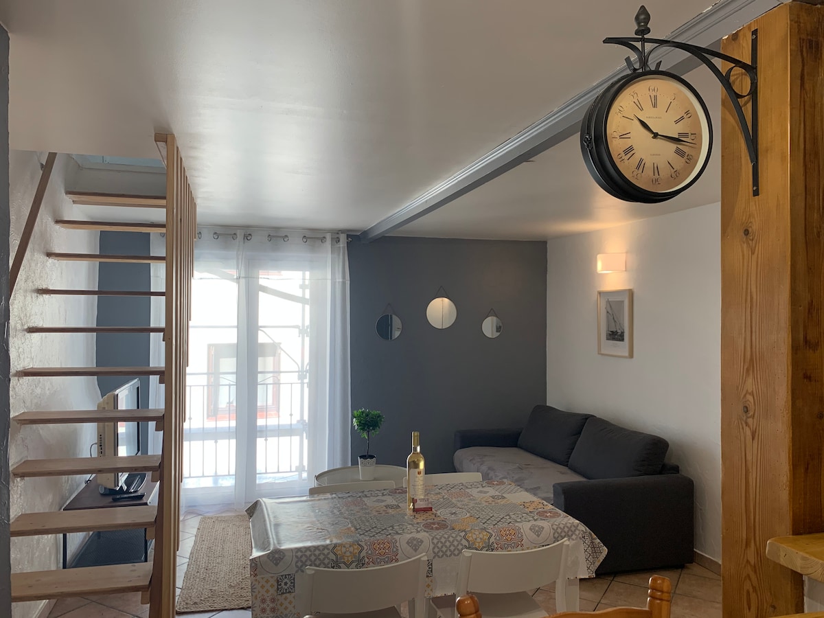 A spacious living area is depicted, featuring a dark gray sofa against a neutral wall. A dining table is positioned in the foreground, adorned with a decorative tablecloth. A distinctive wall clock hangs above, and natural light flows through the double doors leading to a small balcony.
