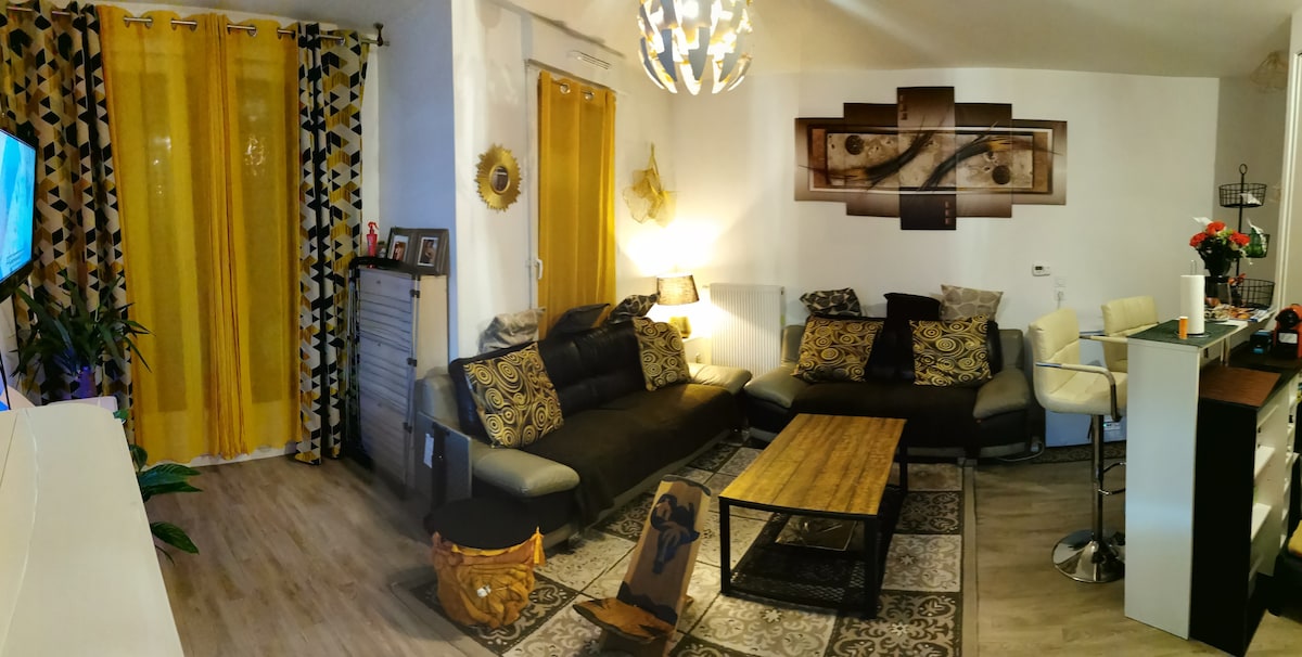 The living area features a spacious black and beige sectional sofa adorned with patterned cushions. A wooden coffee table sits on a decorative area rug, and a modern light fixture hangs from the ceiling. A kitchenette and bar stools are visible in the background, along with bright yellow curtains framing the windows.