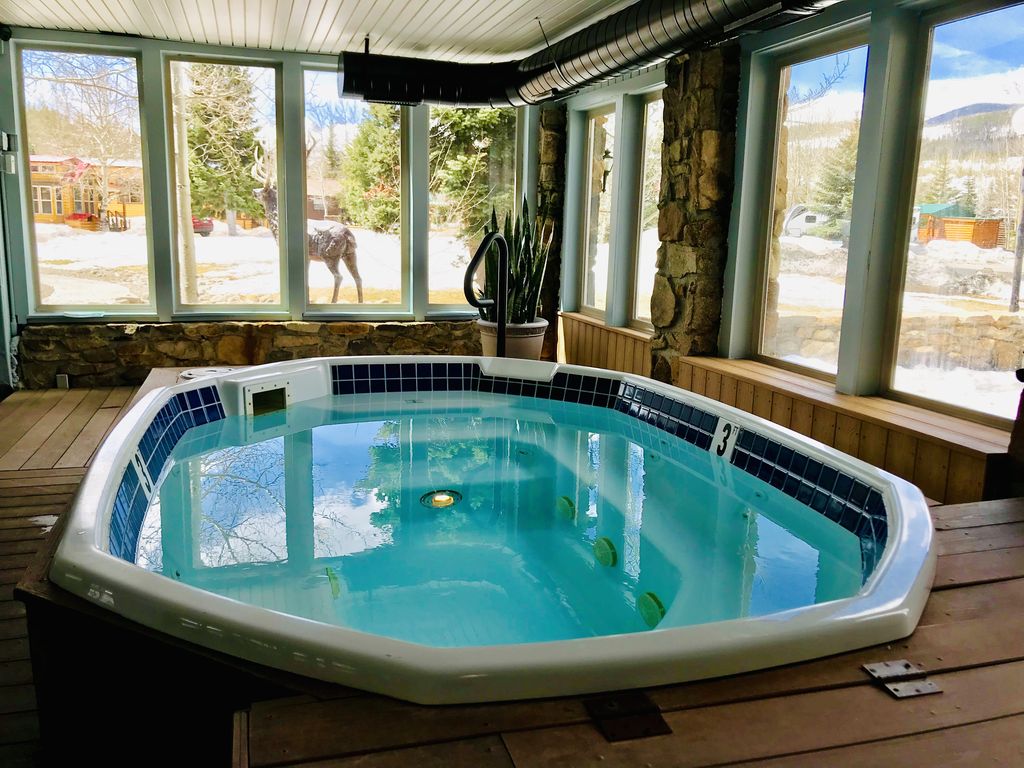 A spacious hot tub is set within a room featuring large windows, allowing for views of the surrounding trees and outdoor landscape. Natural light illuminates the tub and highlights the textured stone walls.