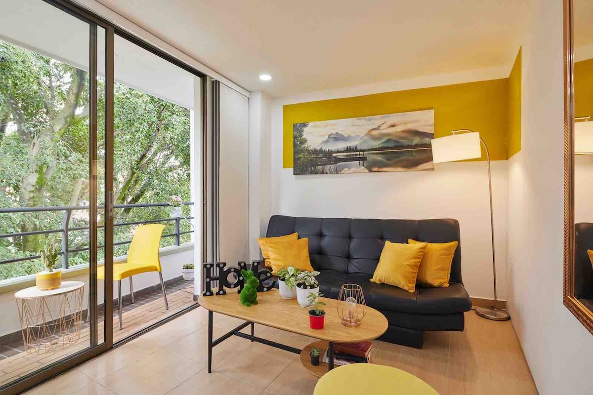 A modern living area features large sliding glass doors that open to a balcony with greenery. A comfortable dark sofa is adorned with yellow pillows, paired with a round wooden coffee table displaying a small plant and decorative items. A floor lamp provides additional lighting in the space.