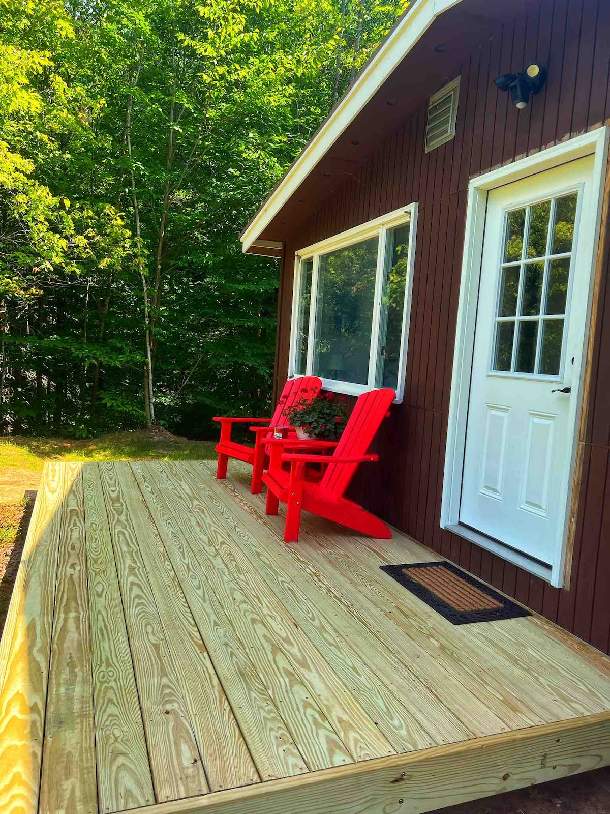 A wooden deck is visible, featuring two bright red chairs that invite relaxation. A door leads into the home, and a window allows natural light to illuminate the space. Lush green foliage surrounds the area, enhancing the serene outdoor setting.