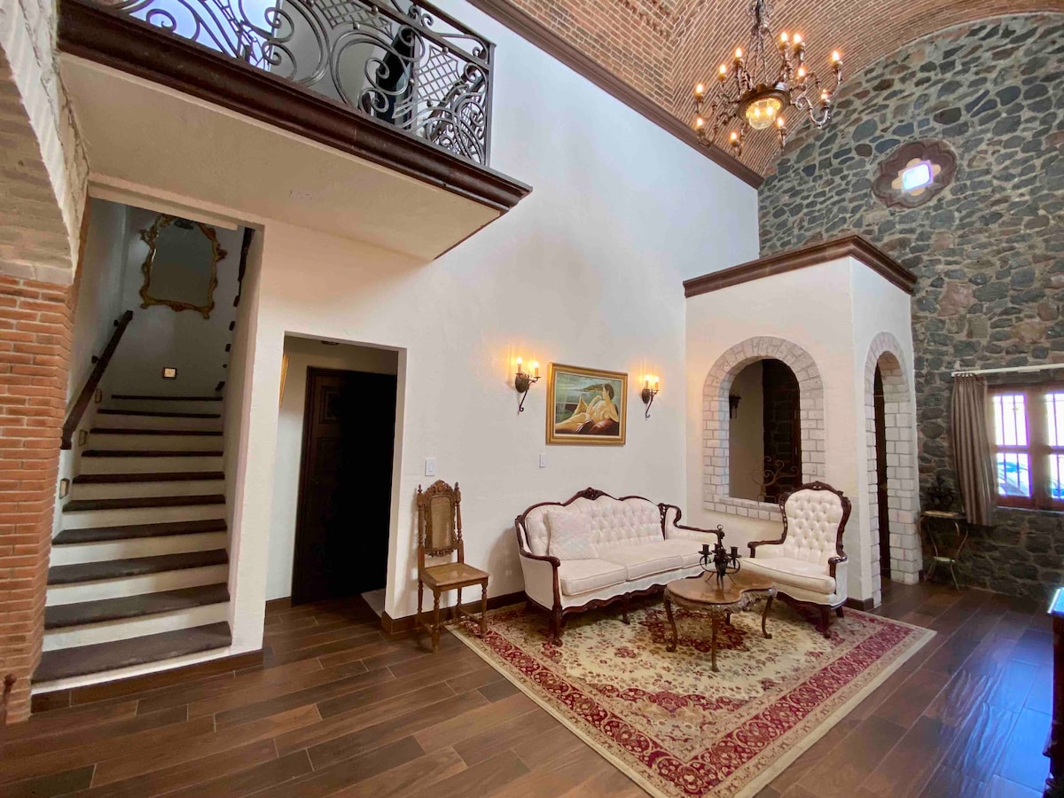A welcoming entrance features elegant seating arranged on a richly patterned area rug. A chandelier hangs above, illuminating the high ceiling with exposed brick and stone walls. A staircase leads to the upper level, while light enters through windows, creating a warm ambiance.