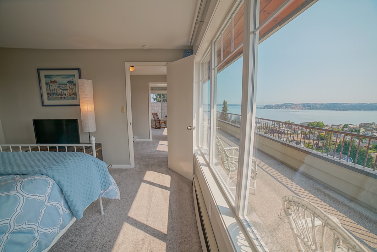 A light-filled bedroom features a spacious bed covered in a soft blue blanket, with expansive windows offering views of the waterfront and distant hills. A television is positioned against the wall, and a door leads to another area of the house.