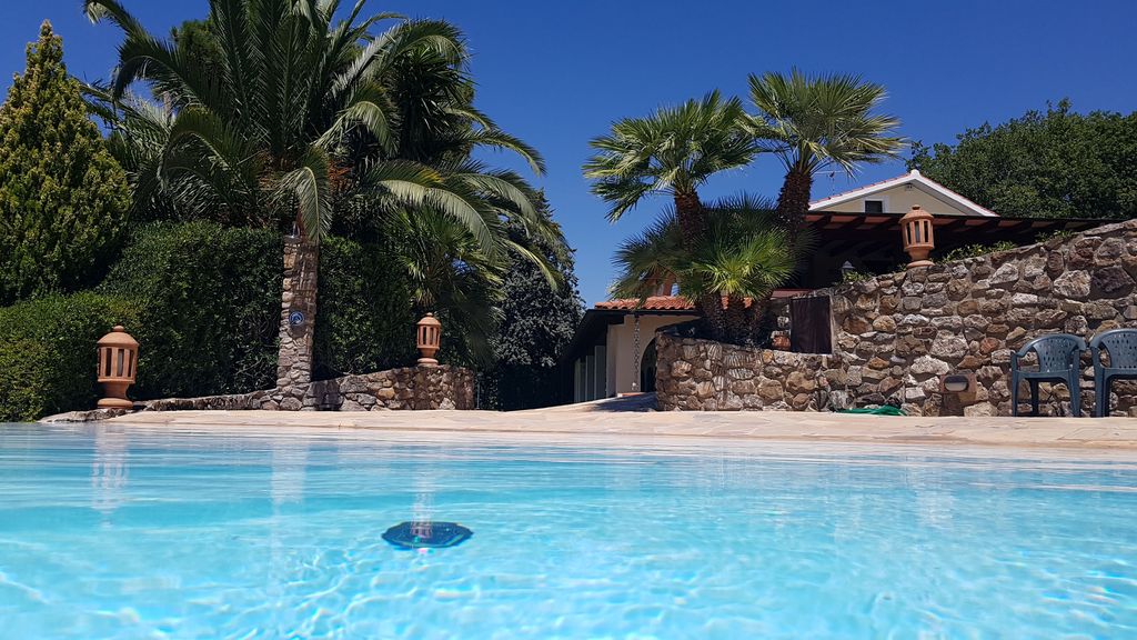 The image captures a tranquil salt-water pool with clear blue water, framed by tropical palm trees. In the background, natural stone walls provide privacy, while decorative lighting fixtures are visible. Two chairs are placed nearby, inviting relaxation in this peaceful outdoor setting.