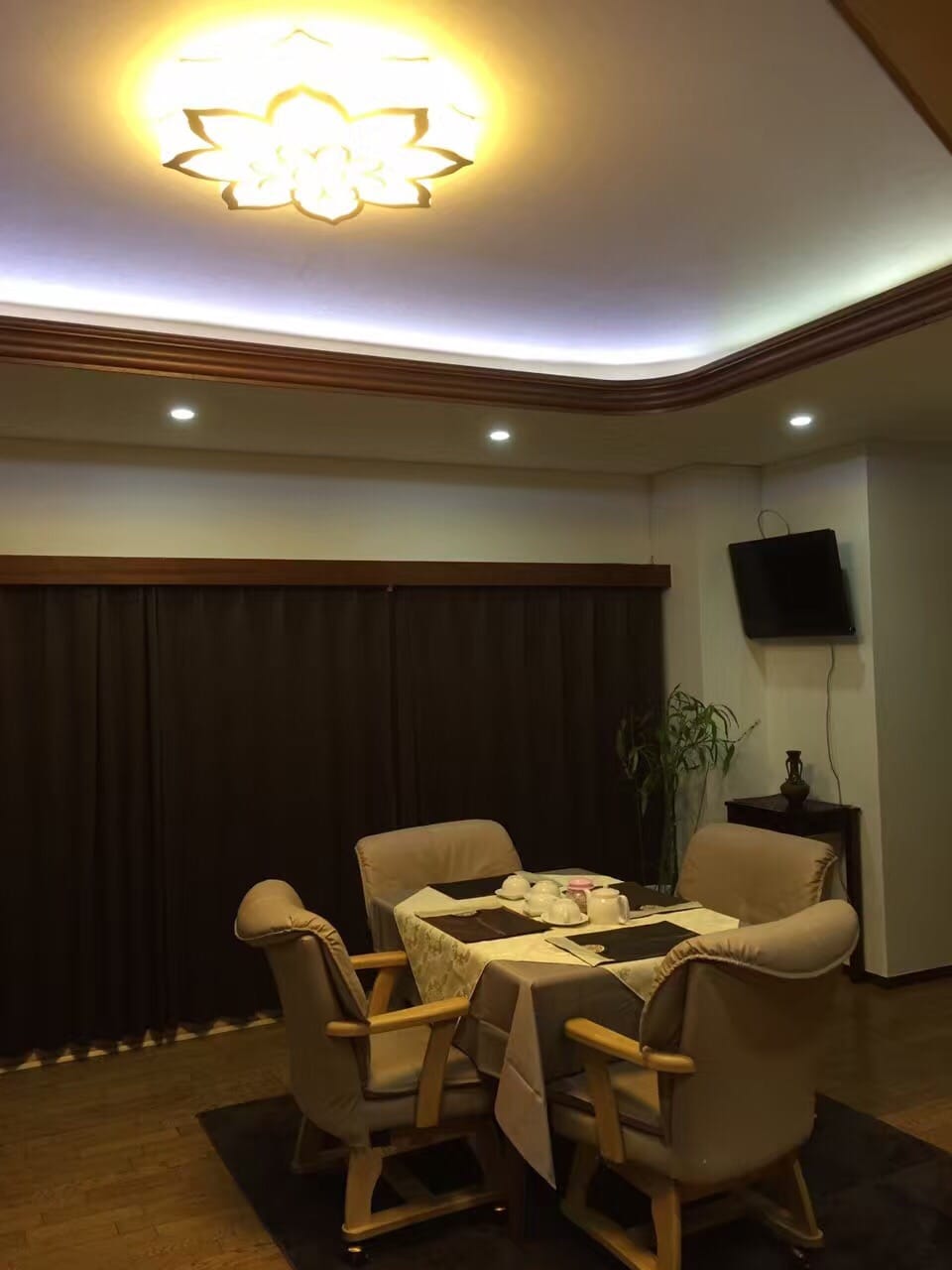 A dining area is presented with a round table set for meals, surrounded by four comfortable chairs. Soft lighting emanates from a decorative ceiling fixture, complemented by recessed lights. Dark curtains and a wall-mounted television enhance the cozy atmosphere of the space.