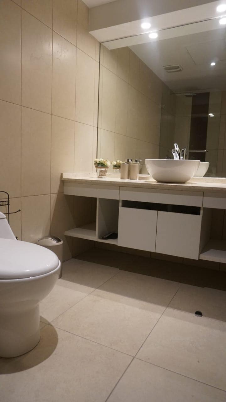 The bathroom features neutral tiled walls with a modern design. A circular sink is positioned on a countertop that offers ample space. Below, a white vanity cabinet provides storage, while a compact toilet is situated to the left. Soft lighting enhances the overall ambiance.