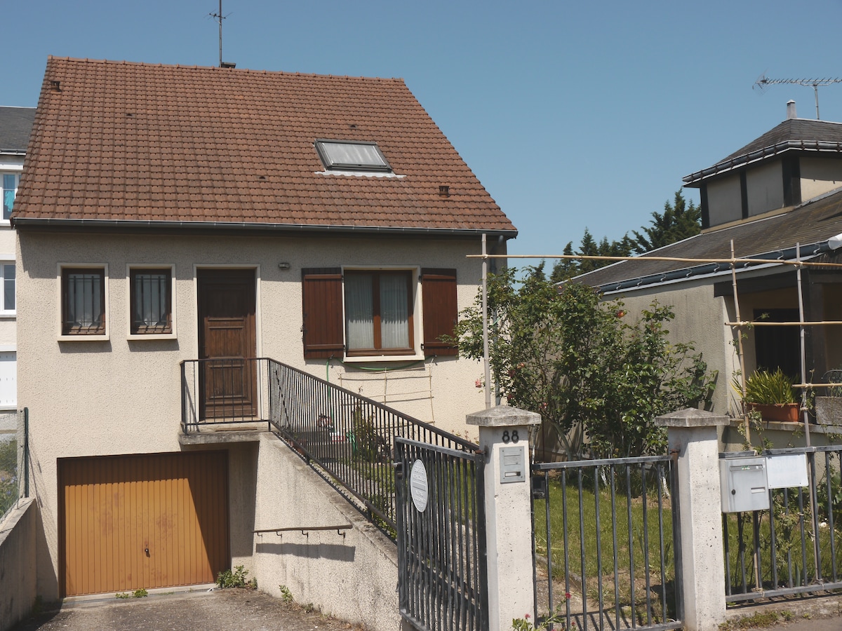 A residential house features a sloped roof with a skylight, framed by brown shutters. The entrance is accessed via a short flight of stairs leading to a wooden door. A small garden area is visible, with greenery surrounding the property.