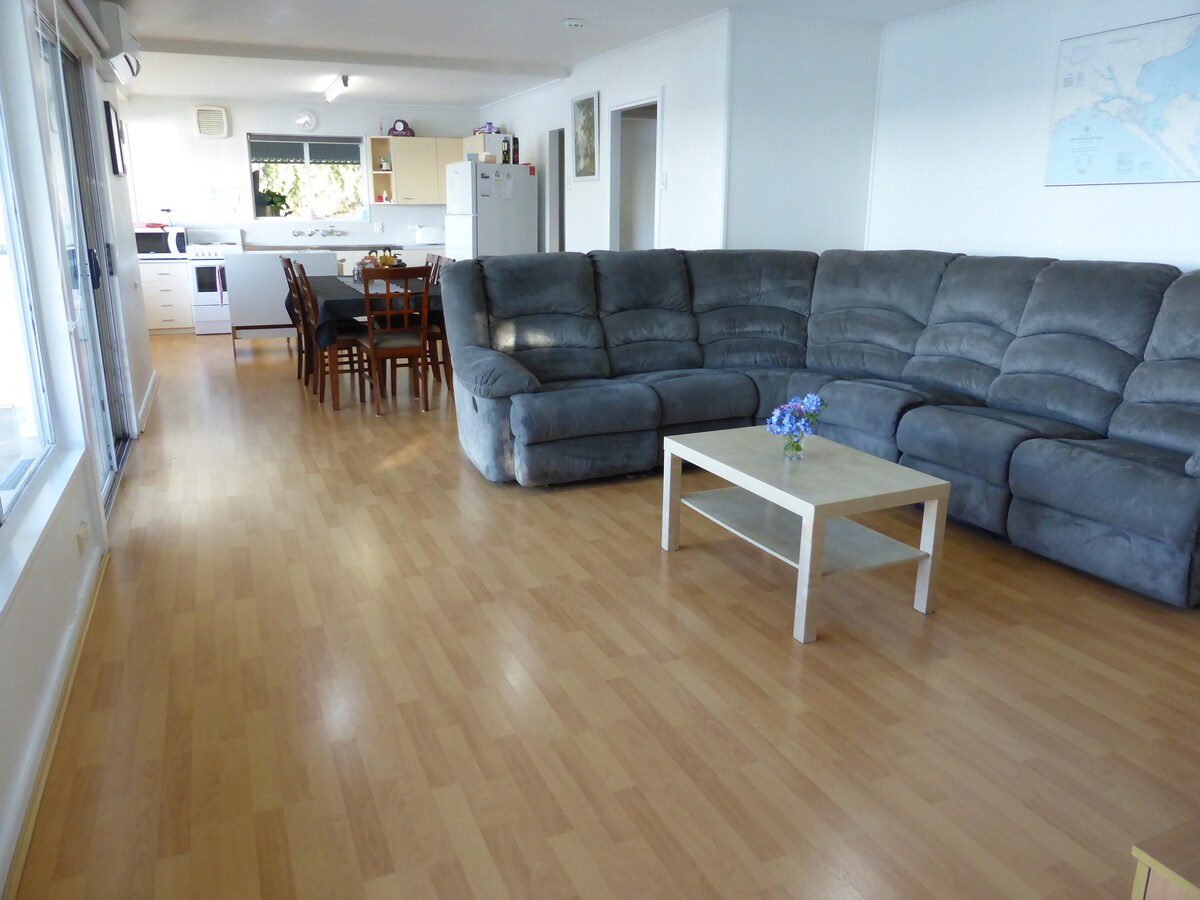 A spacious living area features a large gray sectional sofa, positioned around a light-colored coffee table with a small vase of flowers. Light wood flooring extends throughout the space, leading to a dining area with a wooden table and chairs, visible in the background.
