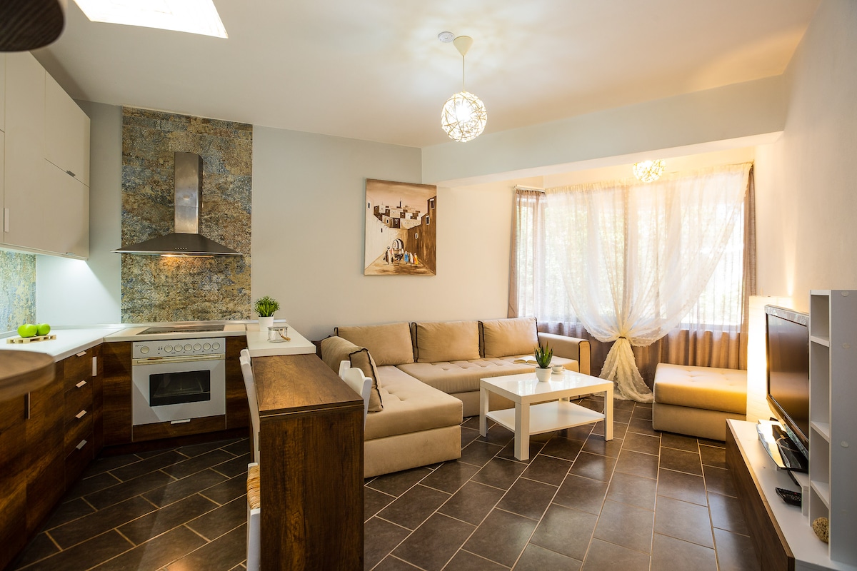 A spacious living area features a modern design with a sectional sofa and a coffee table at its center. Natural light filters through sheer curtains, highlighting the unique wall decor. The kitchen is equipped with contemporary appliances, seamlessly blending with the living space.