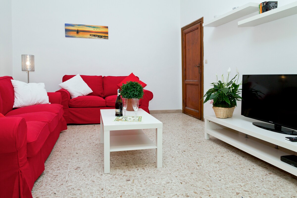 A bright living area is furnished with red sofas and a white coffee table. A television is positioned on a white media console. A decorative plant is placed on the table, complemented by a piece of wall art depicting a serene beach scene.