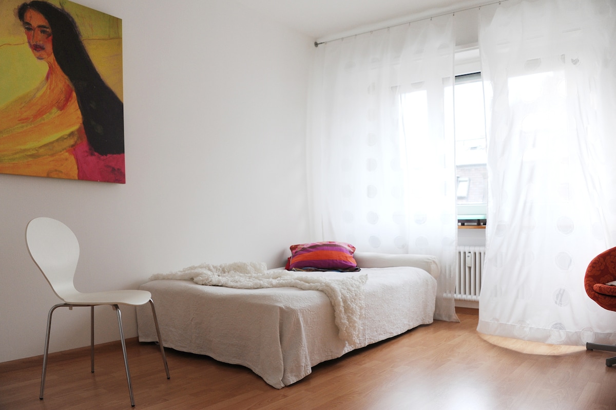 A cozy room features a single bed covered with a light blanket, accompanied by a decorative pillow. A white chair is placed beside the bed. Sheer curtains gently filter light from the window, and a colorful wall art piece adds a touch of vibrancy.