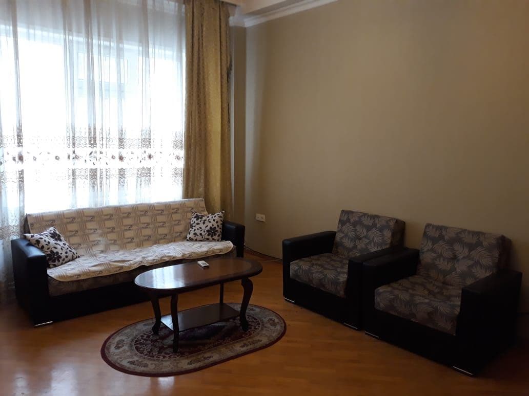 A comfortable living area is presented, featuring a beige sofa with decorative cushions alongside two patterned armchairs. A round coffee table is centered on a decorative rug, while natural light filters through sheer curtains covering large windows.