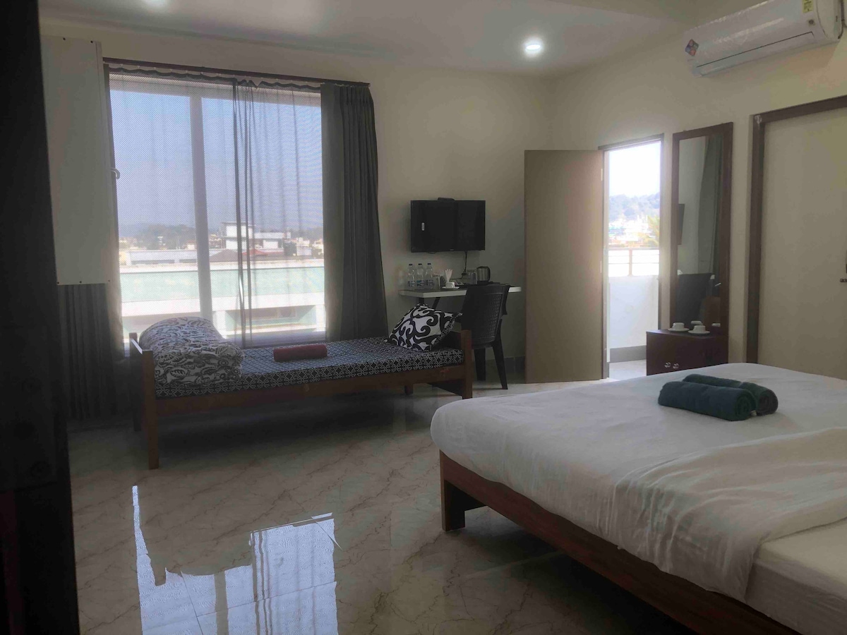A spacious room is equipped with a double bed and a single bed, featuring large windows that allow natural light to fill the space. A small table and chairs are provided near the window. The floor is covered with polished tiles, enhancing the room's brightness.