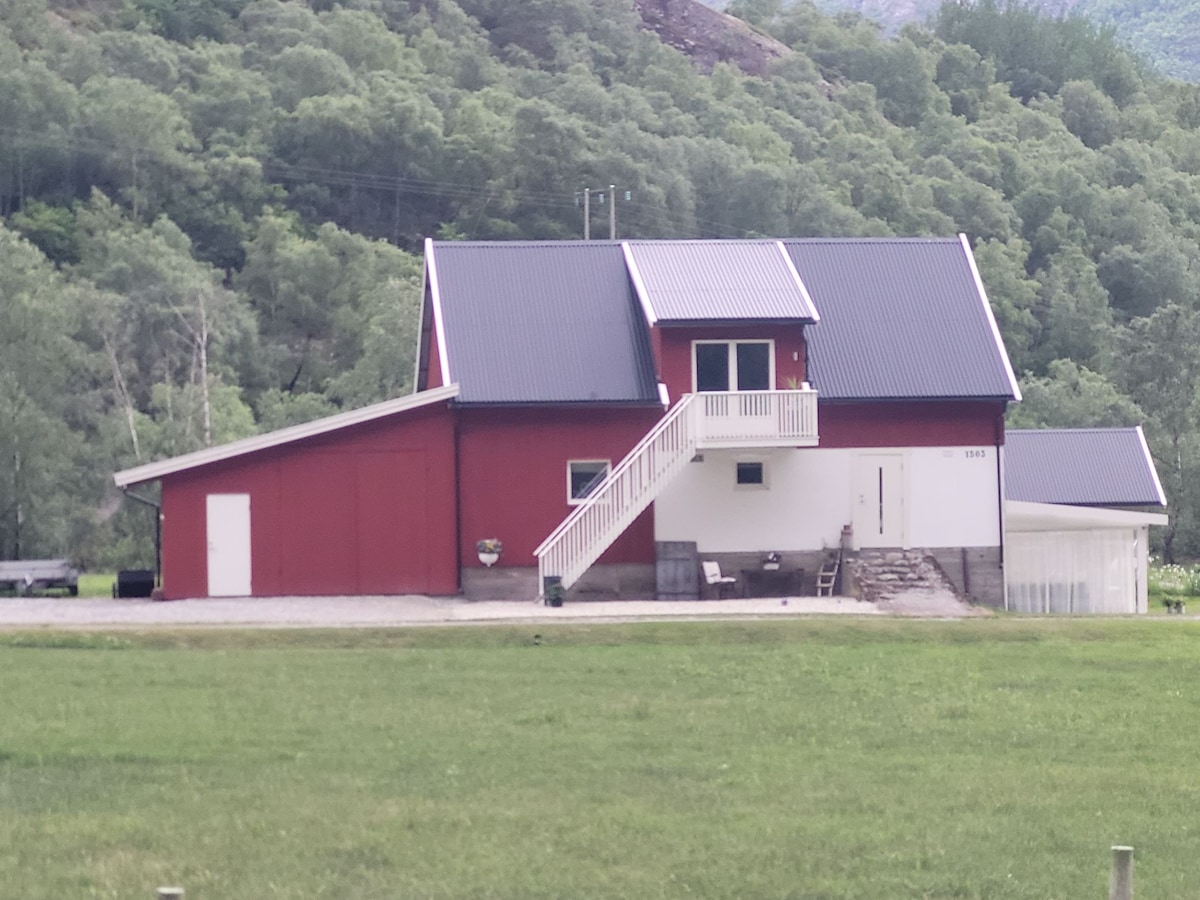 A red, two-story building with a sloped roof is situated amid greenery. A staircase leads to a small balcony on the upper level, while a spacious entryway is visible on the lower level. Surrounding grounds feature a well-kept lawn and nearby trees.