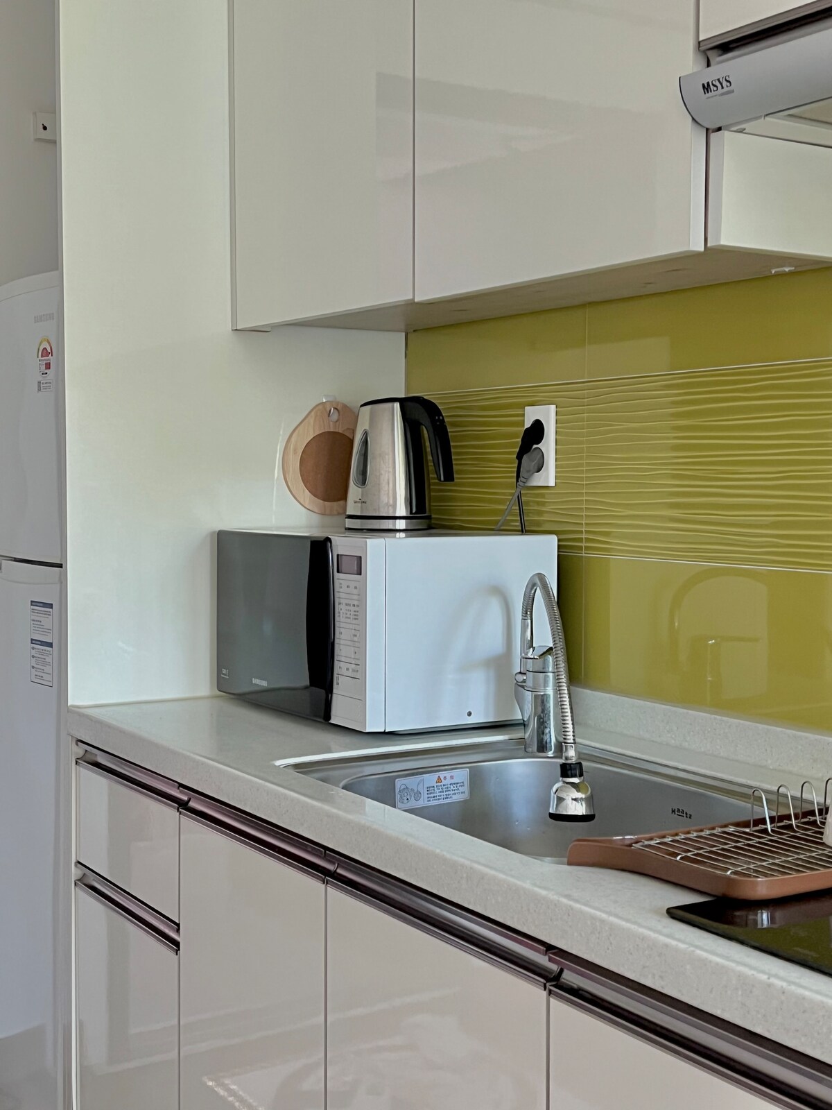 A modern kitchen is equipped with a stainless steel kettle, microwave, and a white countertop. The vibrant green backsplash adds a pop of color. A sink is visible with a dish drying rack nearby, enhancing functionality while maintaining a neat appearance.