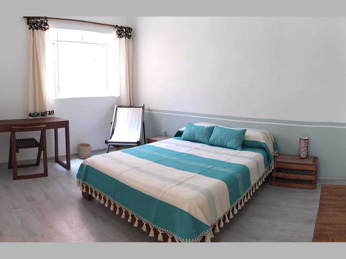 A spacious room features a large bed with a blue and white striped blanket, complemented by two matching pillows. Natural light enters through a window adorned with light curtains. A wooden table and a reclining chair are positioned nearby, creating a functional and inviting space.