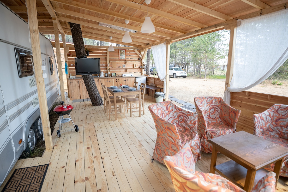 A spacious veranda is showcased, featuring wooden flooring and a seating area with vibrant patterned chairs. A dining table is positioned nearby, while a television is mounted on a wooden wall. A kitchenette, equipped with essential appliances, is visible to the side.