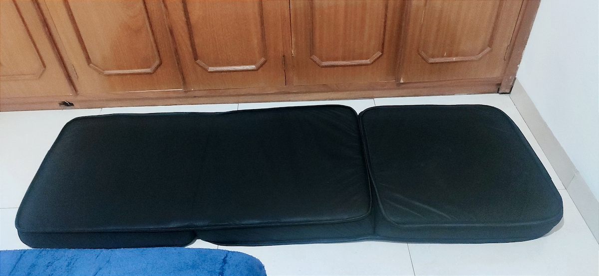 A black foam mattress pad is displayed on a tiled floor, featuring a flat surface with stitched edges. The background consists of wooden cabinetry, enhancing the sense of space and practicality.