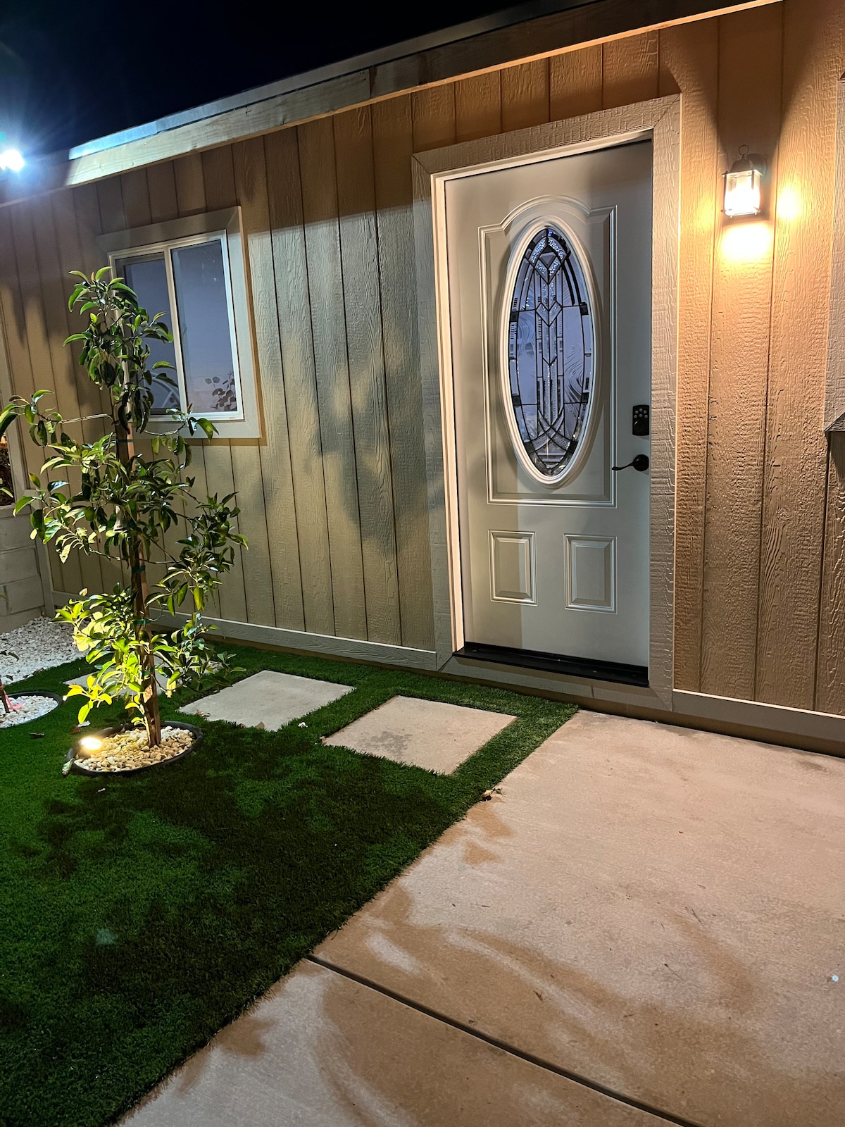 The entrance to the guest house features a well-lit doorway with an oval window and a decorative light fixture. A small tree is positioned beside the entrance, enhancing the welcoming ambiance. Artificial grass provides a neat, green border alongside the decorative pathway leading to the door.