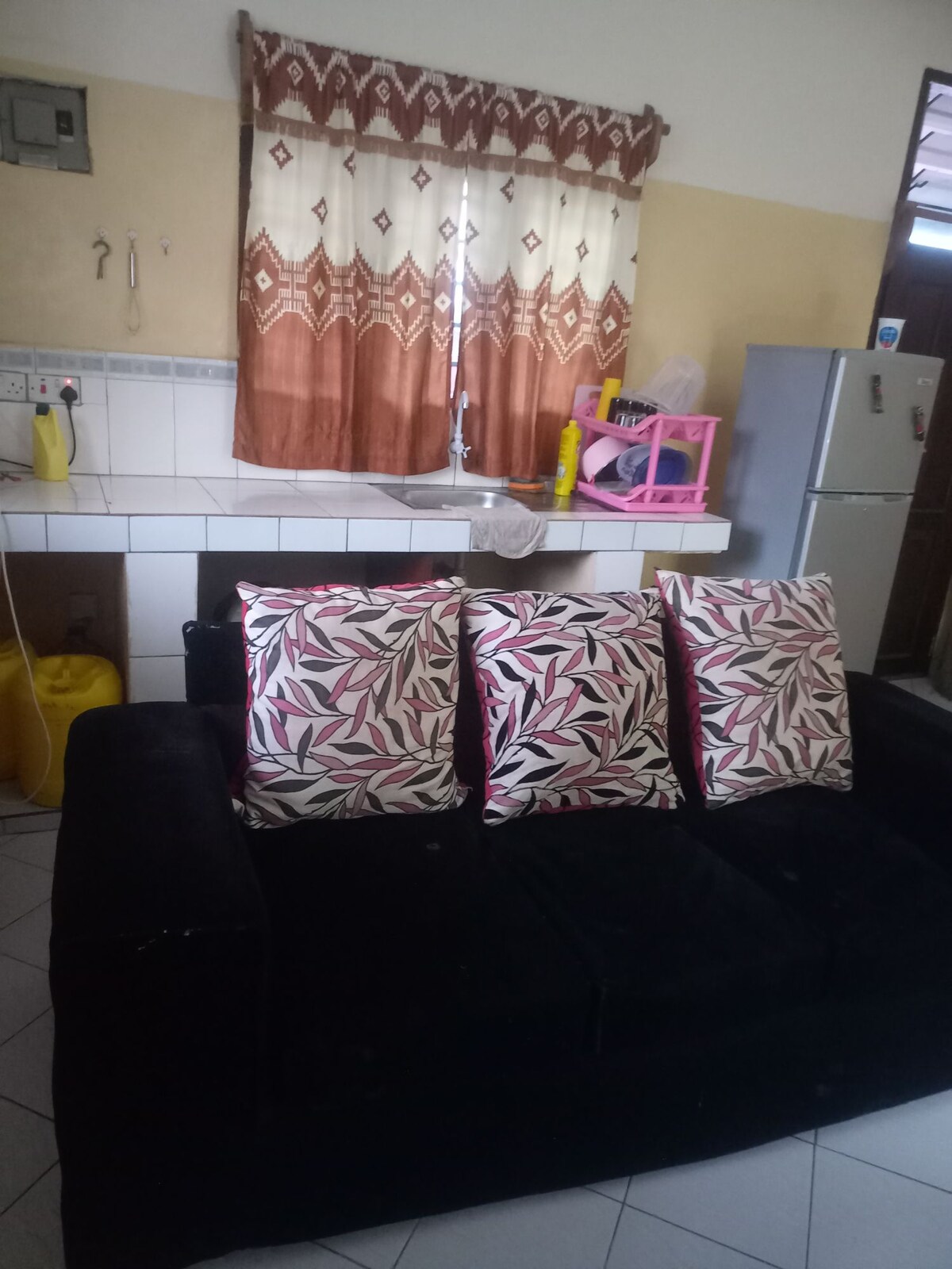 A dark sofa is adorned with three cushions featuring vibrant leaf patterns. Behind the sofa, a kitchen area is visible, showcasing a sink and a fridge. Natural light filters through a patterned curtain, adding warmth to the space.
