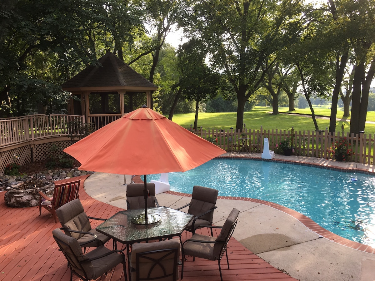 An outdoor area features a refreshing pool surrounded by a wooden deck. A gazebo is positioned nearby, offering shaded seating under a vibrant orange umbrella. Lush greenery and a fenced area enhance the tranquil ambiance of this recreational space.