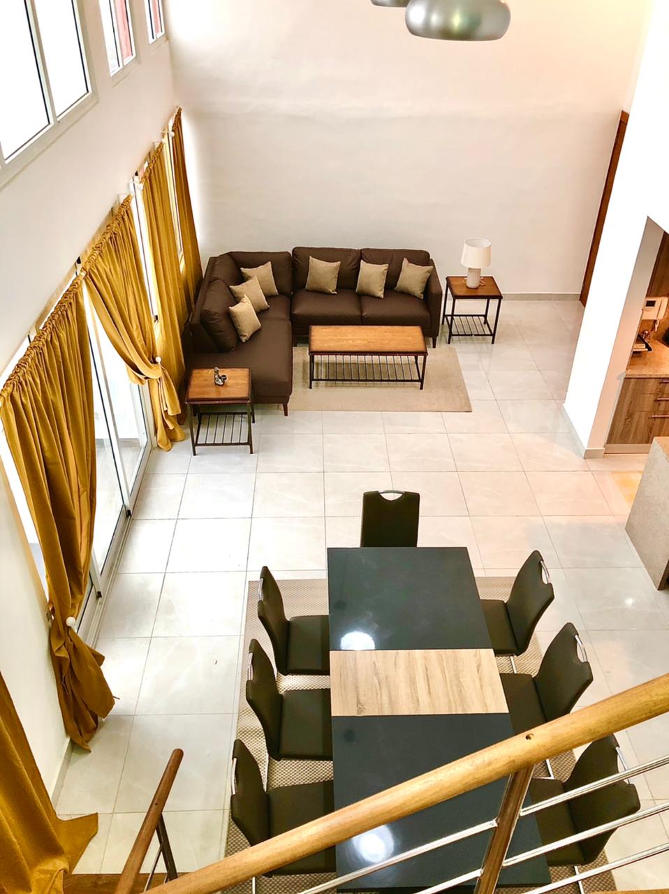 A spacious living and dining area is visible, featuring a large brown sectional sofa adorned with decorative cushions. A wooden coffee table sits in front, complemented by a dining table with six dark chairs. Large windows provide ample natural light, framed by soft, golden curtains.