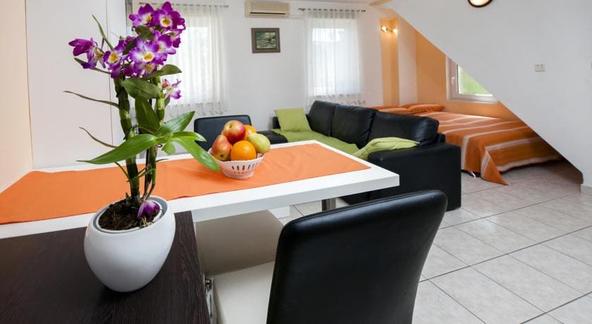 A welcoming living space features a contemporary dining table with a bowl of fresh fruit and a decorative plant. The area is furnished with a black leather sofa and a bright green couch, with sunlight filtering through sheer white curtains, creating a cheerful ambiance.