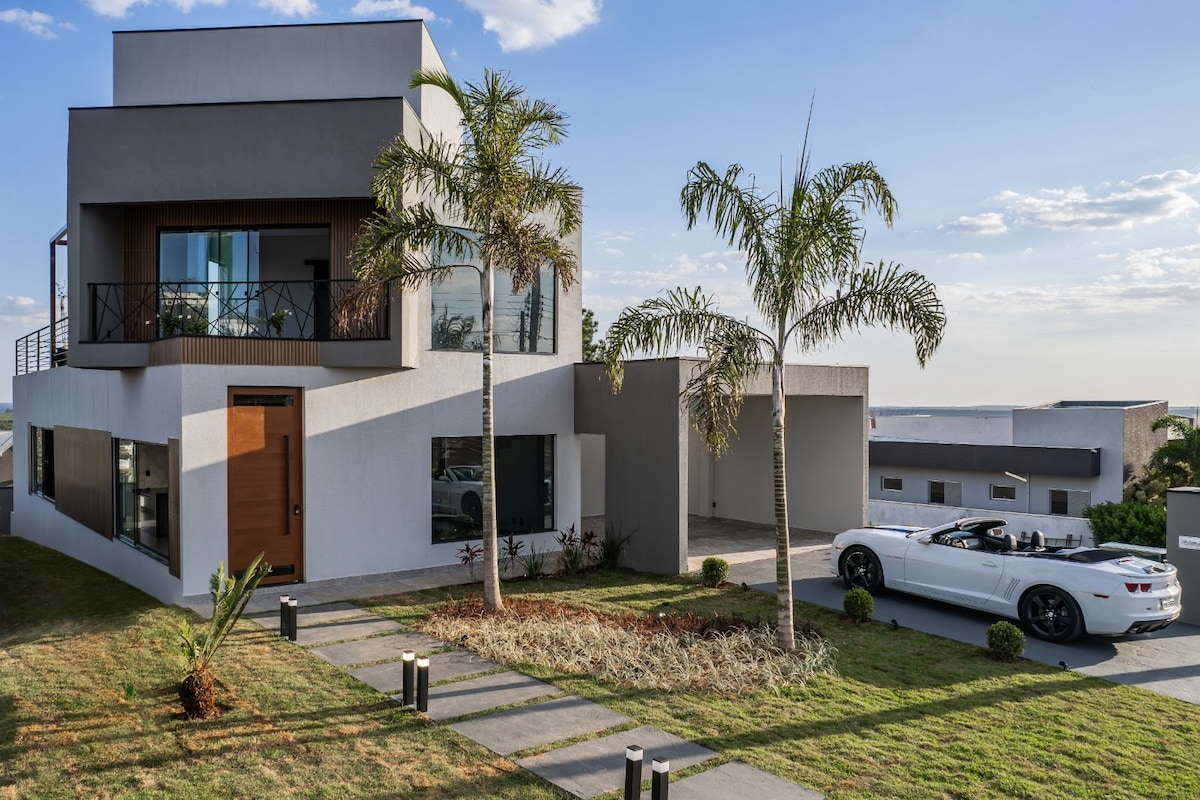 A modern two-story house is presented with large glass windows and a contemporary design. A white convertible car is parked in front, on a landscaped lawn featuring palm trees and a stone pathway leading to the entrance.
