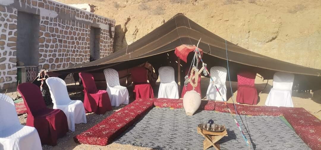 A spacious outdoor seating area is covered by a large, dark canopy. White and maroon chairs are positioned around a traditional woven rug, while a low wooden table is placed in the center. Decorative items, including a stringed instrument, are visible, reflecting local culture.