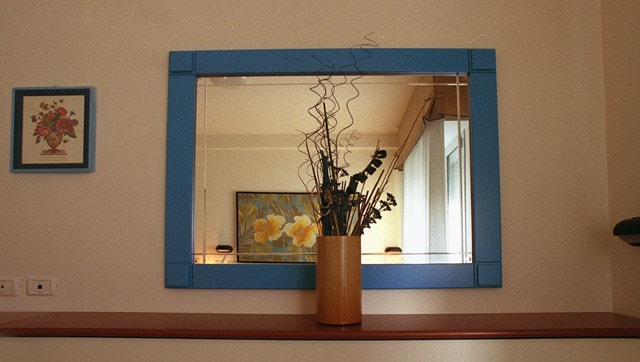 A large mirror with a blue frame is positioned above a wooden shelf. A round vase containing decorative twigs and leaves is centered under the mirror. A framed floral artwork hangs on the wall to the left, complementing the overall aesthetic.