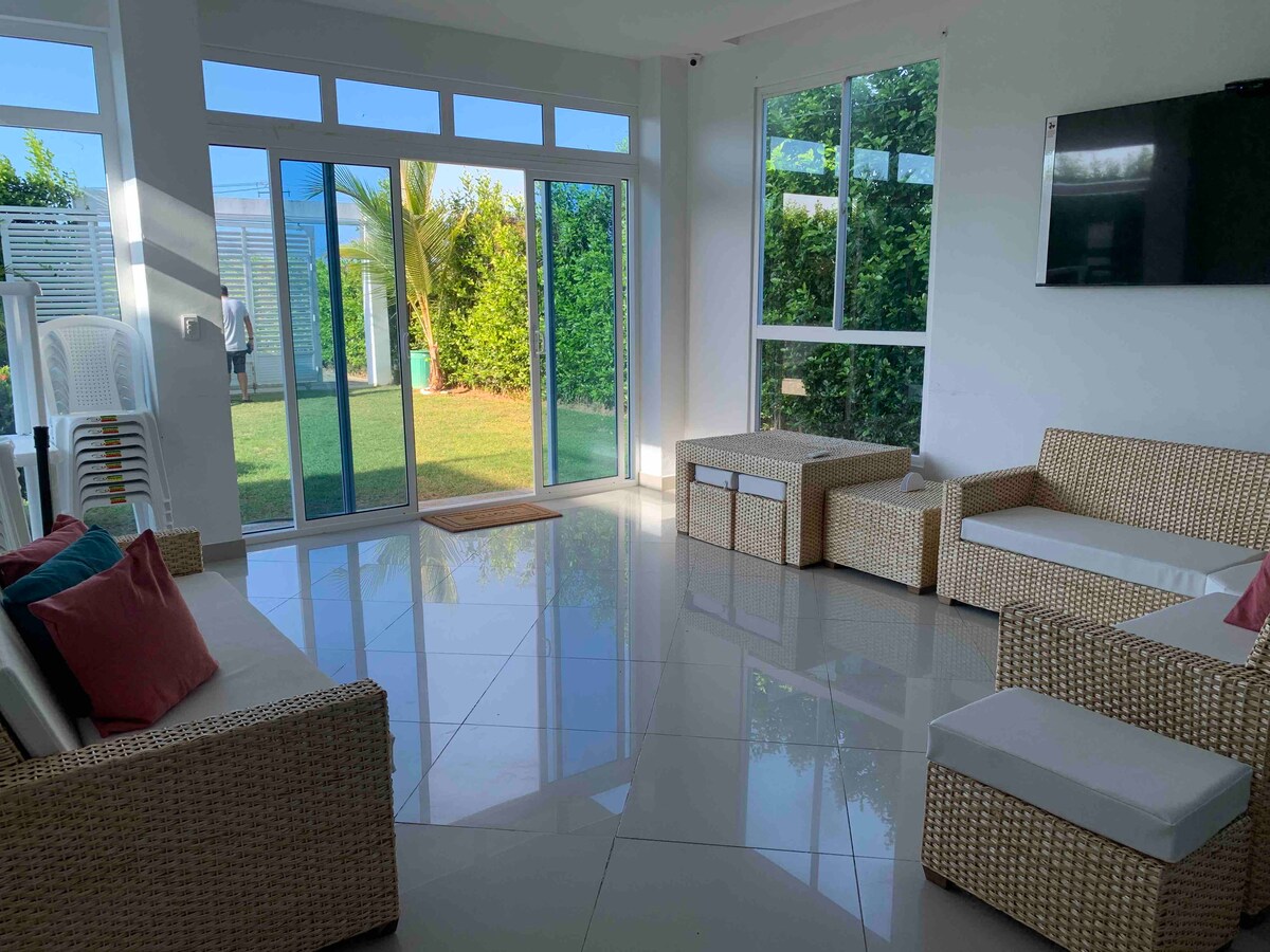 A bright and airy living space features large glass doors that open to a well-maintained garden. Comfortable seating is arranged on glossy tiled flooring, with soft cushions adding subtle color. A wall-mounted television is visible, enhancing the space's functional appeal.