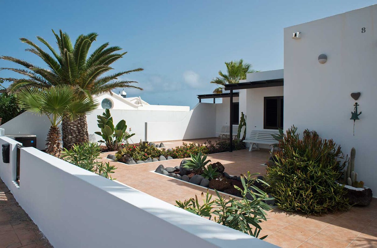 A garden area is depicted, featuring a variety of lush plants and palm trees surrounding a landscaped space. A seating area is present, and natural stone elements enhance the outdoor environment. The white walls create a bright contrast against the greenery and blue sky.