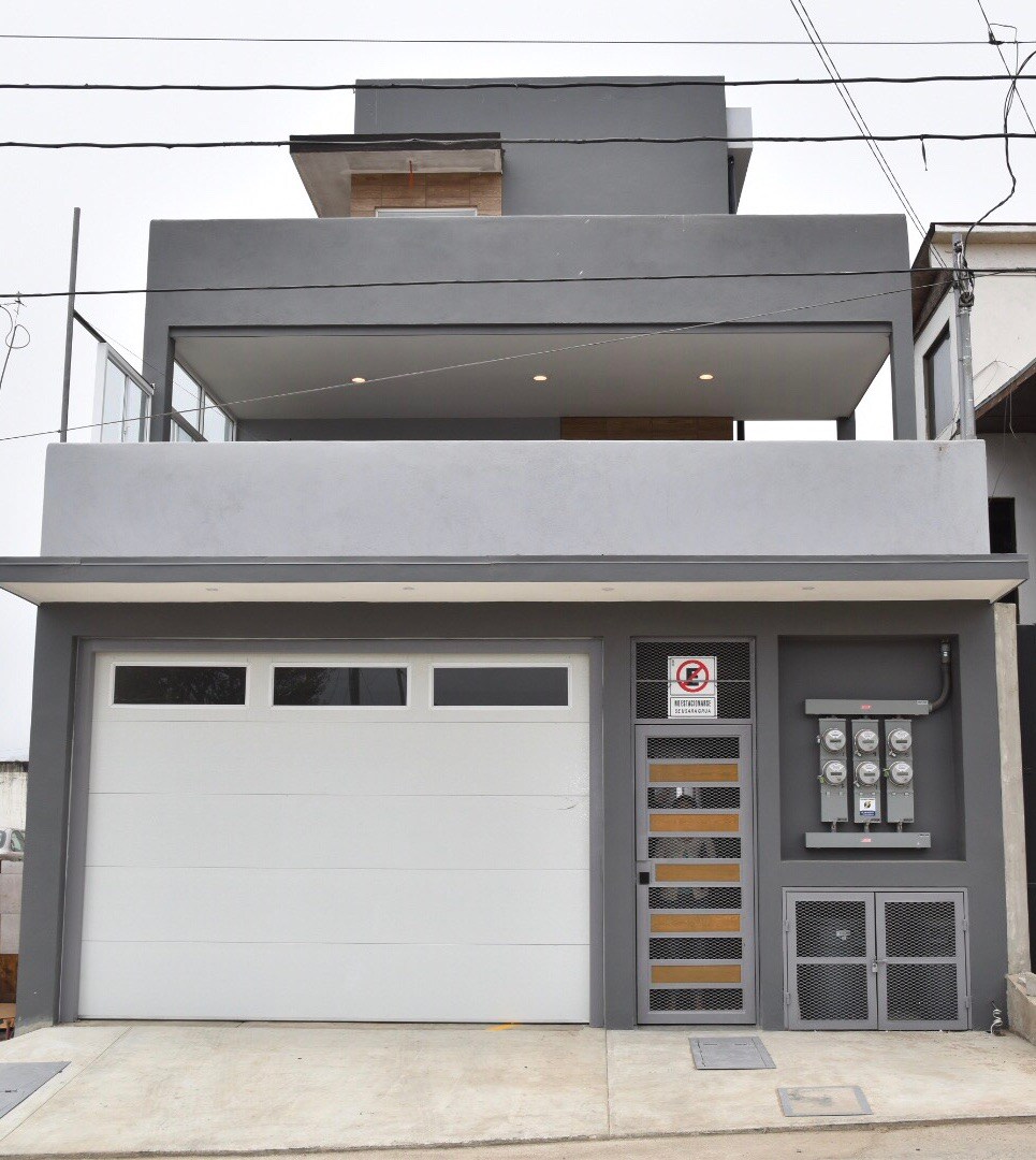 A modern two-story building with a sleek gray exterior. The entrance features a garage with a white door and utility meters alongside. An upper balcony is visible, complemented by large, well-distributed windows that enhance natural light throughout the structure.