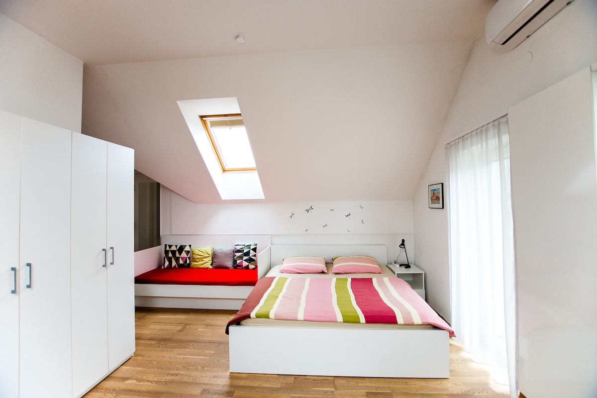 A bright sleeping area features a spacious bed adorned with striped bedding and a cozy seating nook with colorful cushions. A window with a skylight invites natural light, and modern white cabinetry completes the airy atmosphere.