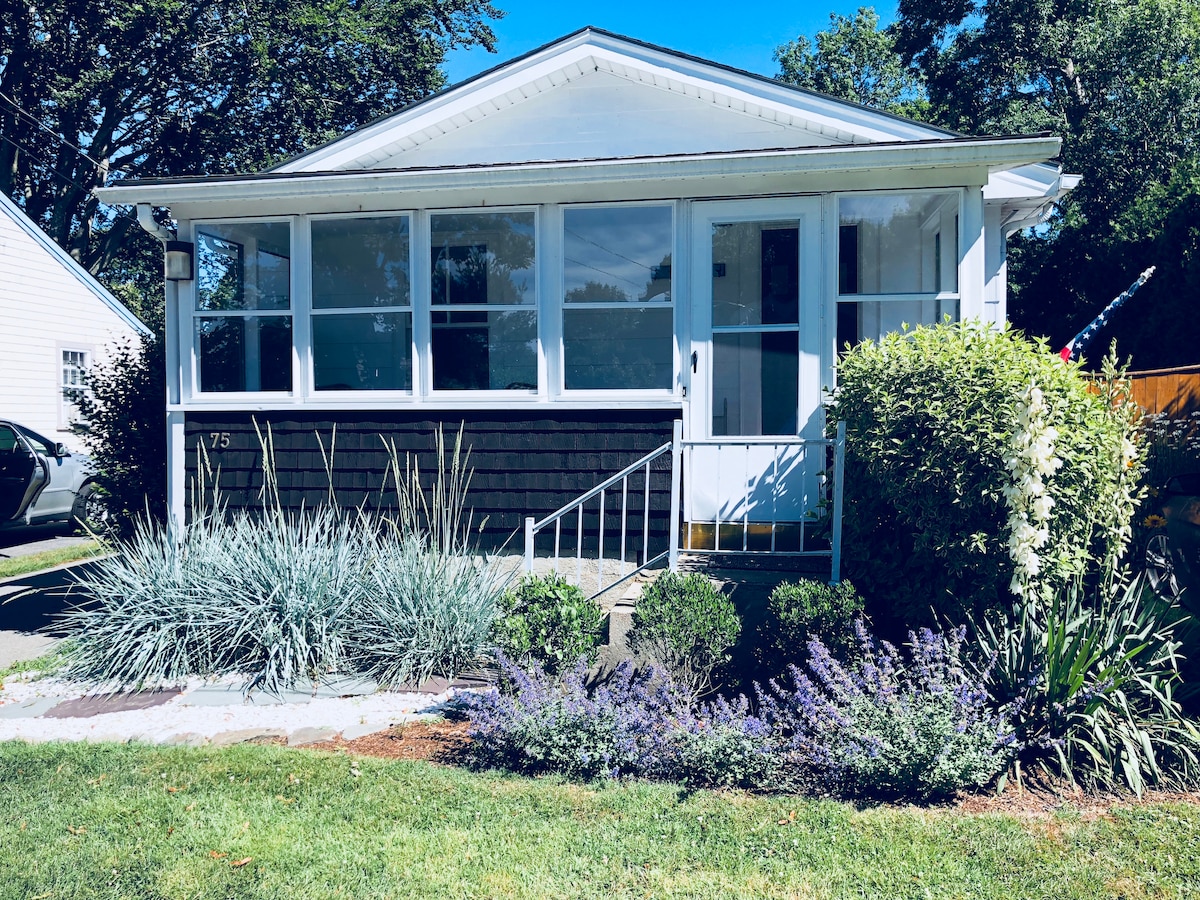 A charming front view of the house features large windows allowing natural light. Lush green shrubs and colorful flowers frame the entrance, while a well-maintained lawn enhances the inviting exterior. The porch steps lead to the main door, creating an appealing entryway.