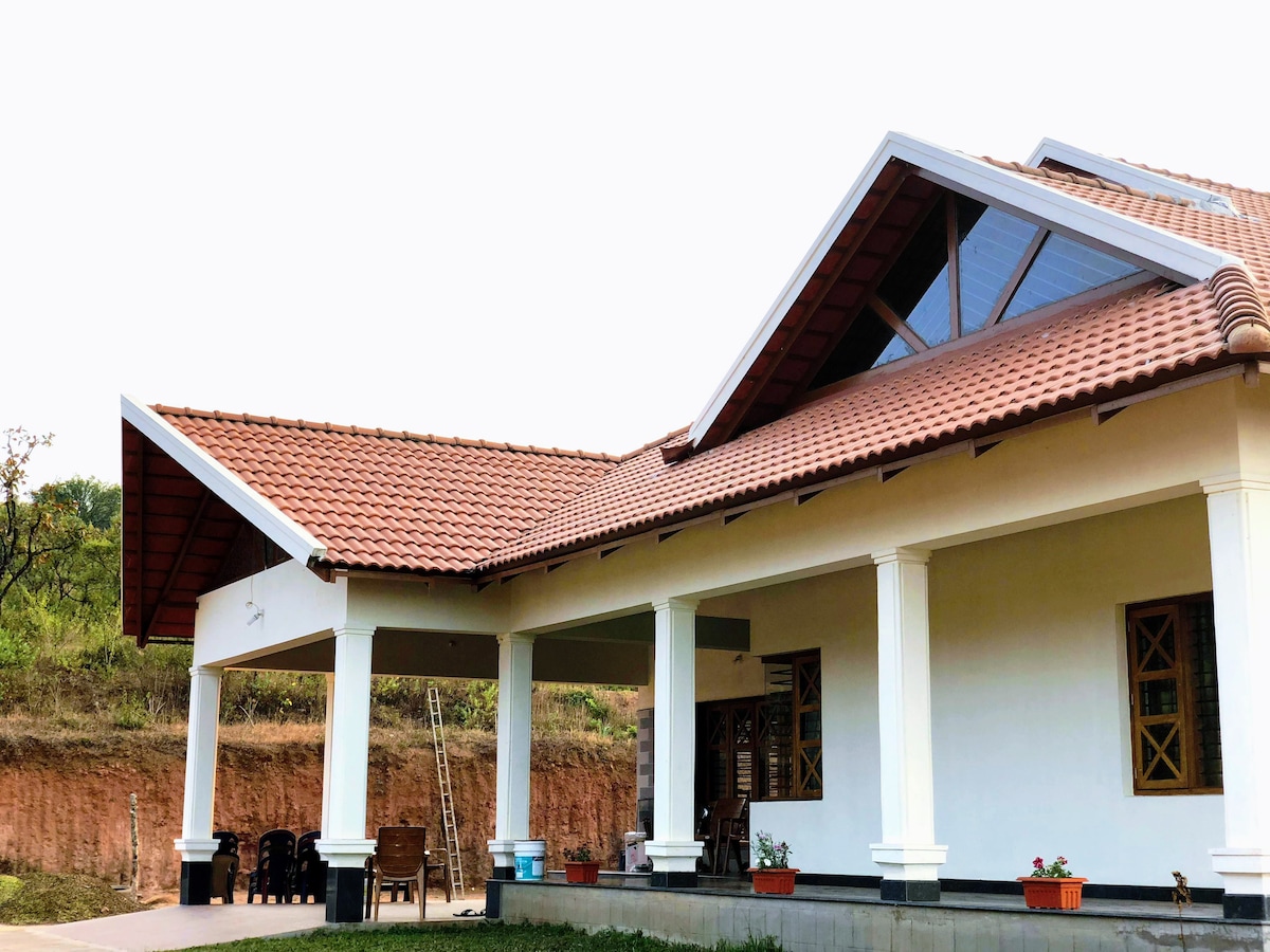 The exterior of a spacious homestay showcases a traditional architecture with a tiled roof and large pillars. An open area is visible, featuring potted plants and seating arrangements. A sloped hill can be seen in the background, providing a natural backdrop.
