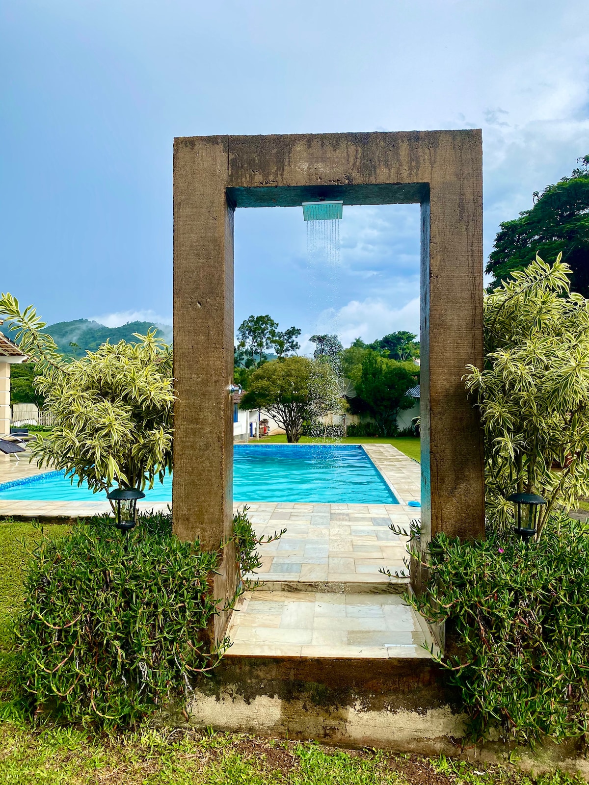 An archway framed by lush greenery leads to a serene pool area. The clear blue water of the pool reflects the surrounding landscape, while a soft cascade of water flows in the background. The atmosphere is peaceful, inviting relaxation and leisure.