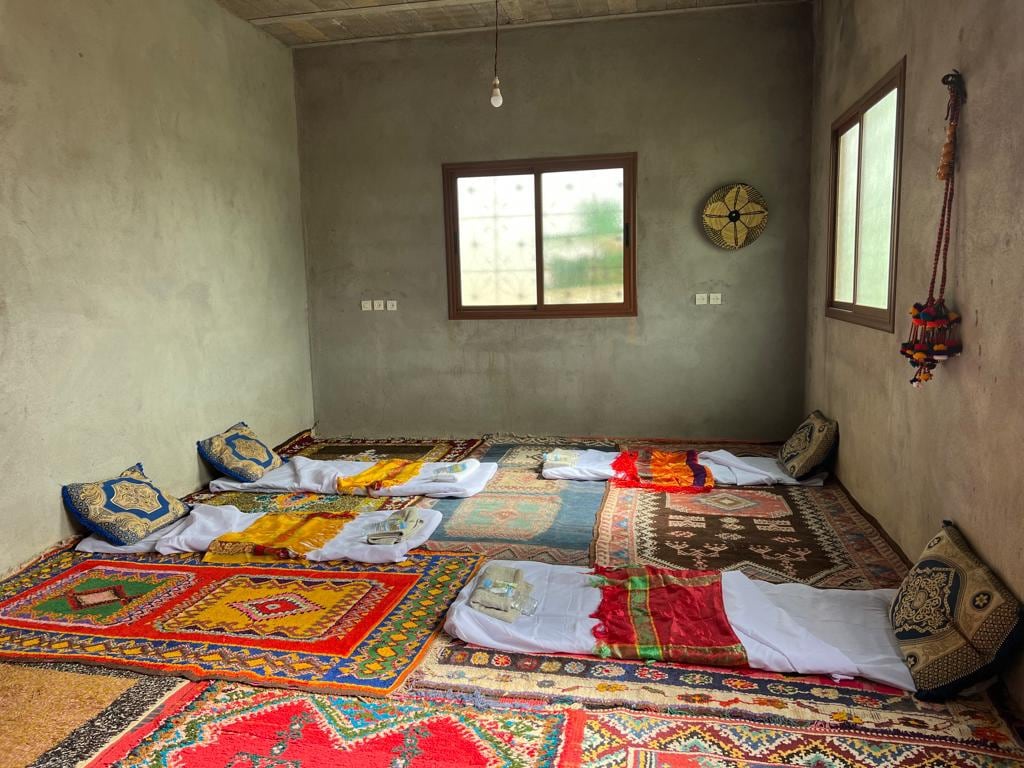 A simple sleeping area is presented, featuring colorful woven carpets and multiple laid-out bedding arrangements situated on the floor. Walls are finished in a muted shade, and natural light enters through a large window, enhancing the spaciousness of the room.